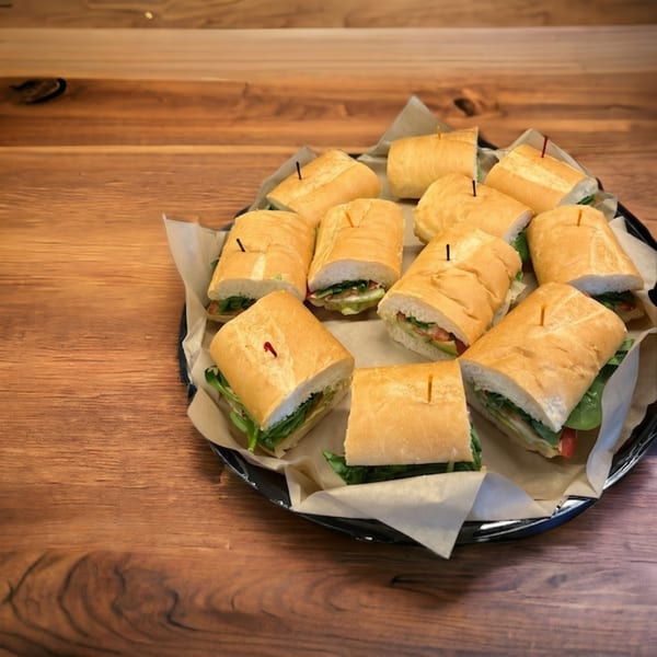 A black platter filled with numerous baguette sandwiches, each cut in half and wrapped in parchment paper.